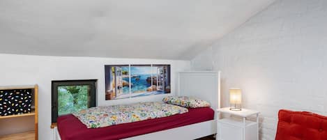 Ein Schlafzimmer mit einem Bett, einem Nachttisch, einem Sessel, einem Regal und einer Wand mit einem Bild.