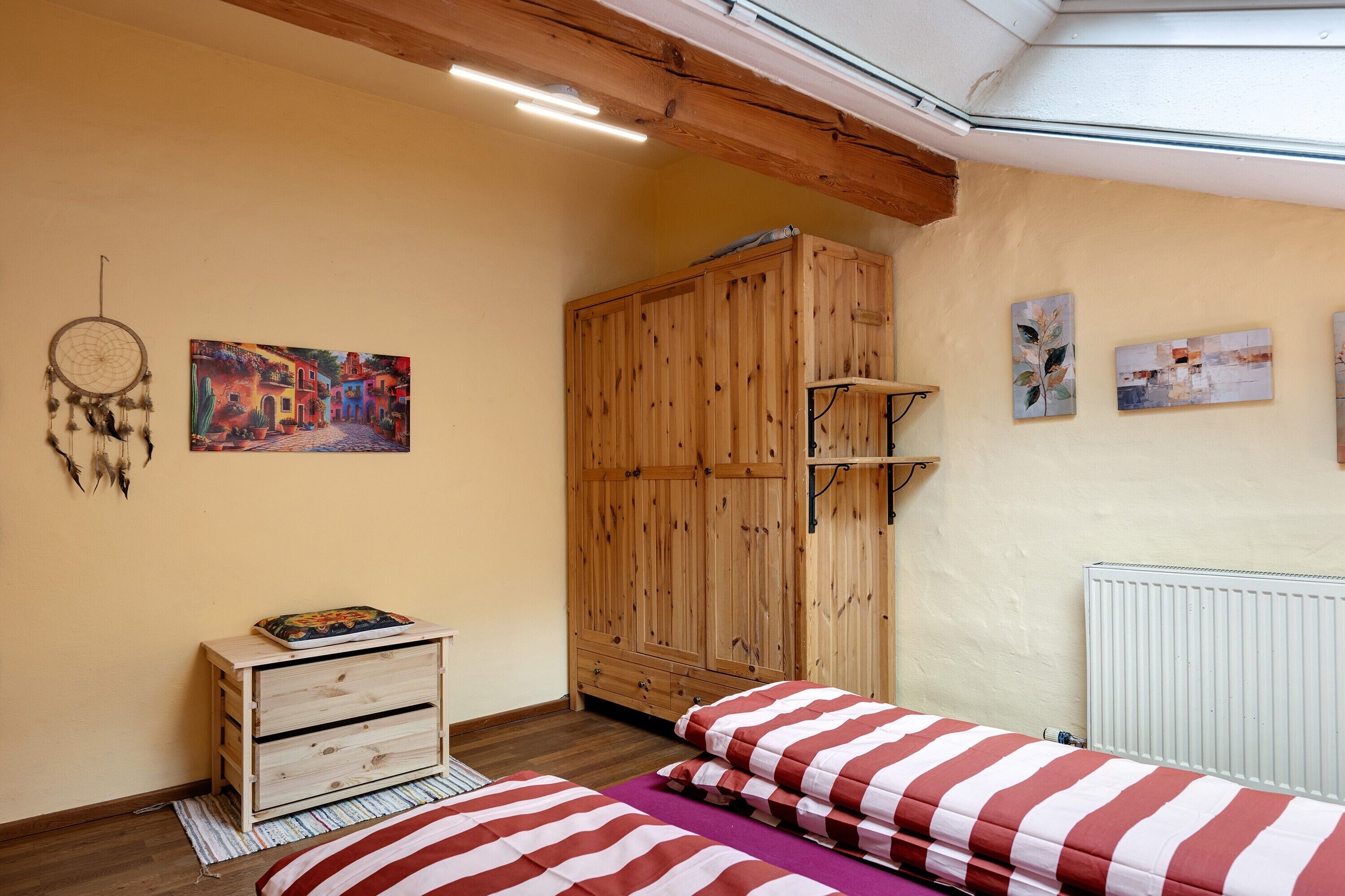 Ein Schlafzimmer mit Holzschrank, einem Bett mit gestreifter Bettwäsche, einem Traumfänger und zwei Wandbildern.