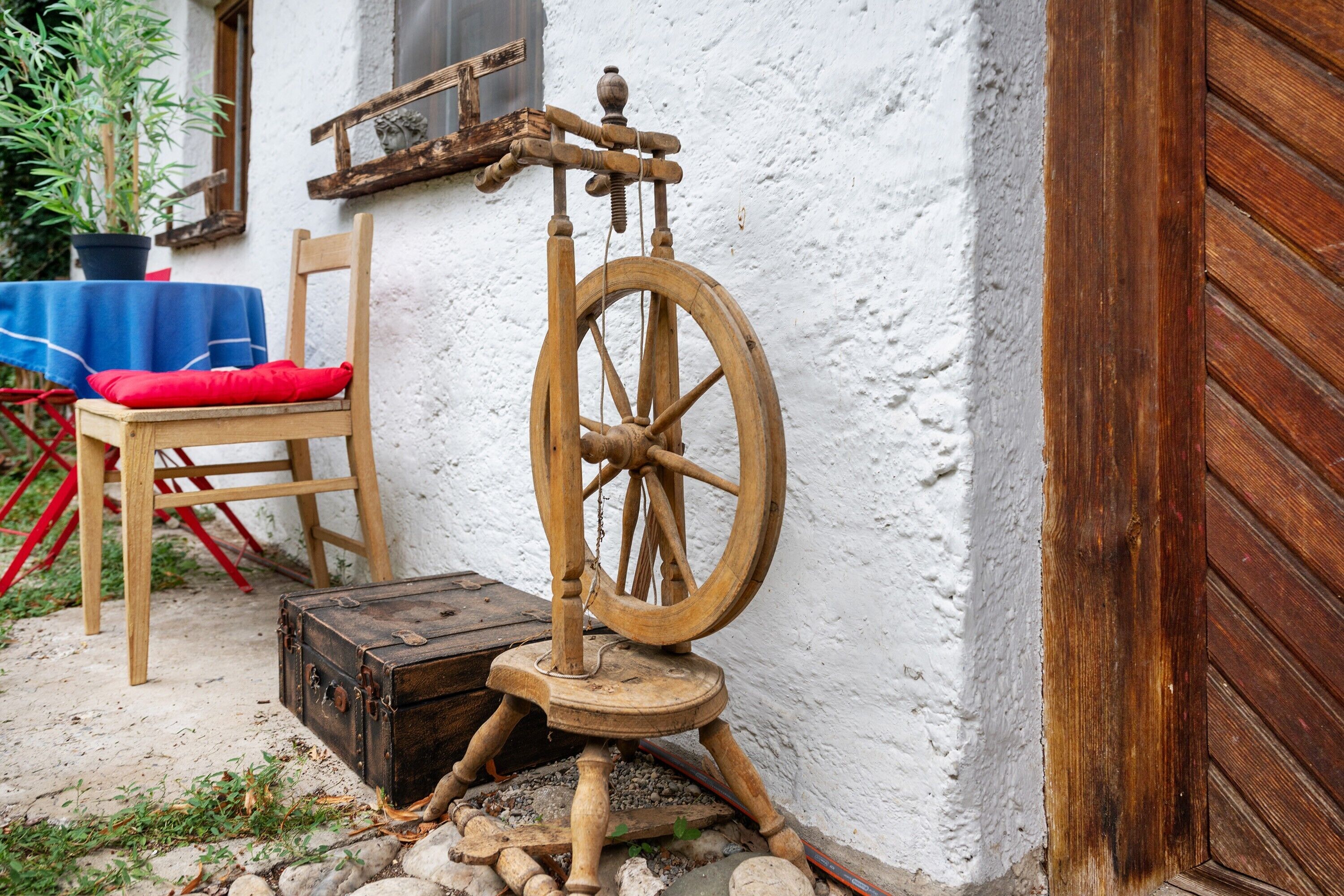 A rustic outdoor setting with a wooden spinning wheel, a wooden chair with a red cushion, and a small wooden chest.