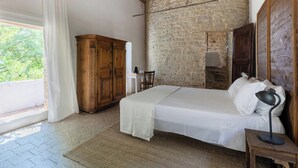 A bedroom with a stone wall, a wooden wardrobe, a bed with white bedding, and a desk with a lamp.