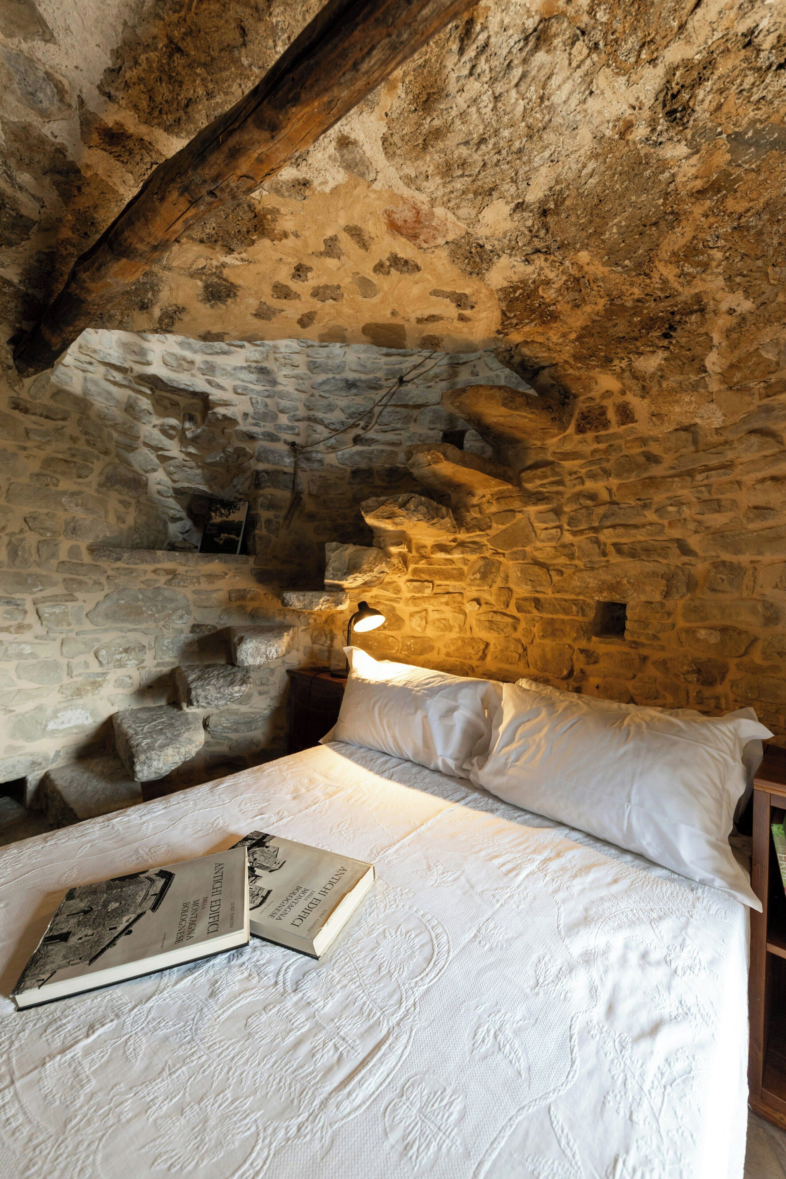 A stone-walled room with a bed, a book, and a lamp.
