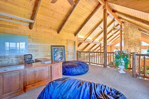 Um quarto espaçoso com teto de madeira alto, uma escrivaninha, uma cadeira, um grande puff e vista para o exterior.