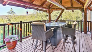 A covered outdoor deck with a table and chairs, surrounded by a wooden railing, overlooking a forested area. - Luxe Prescott Natl Forest Home w/ Huge Deck (Prescott)
