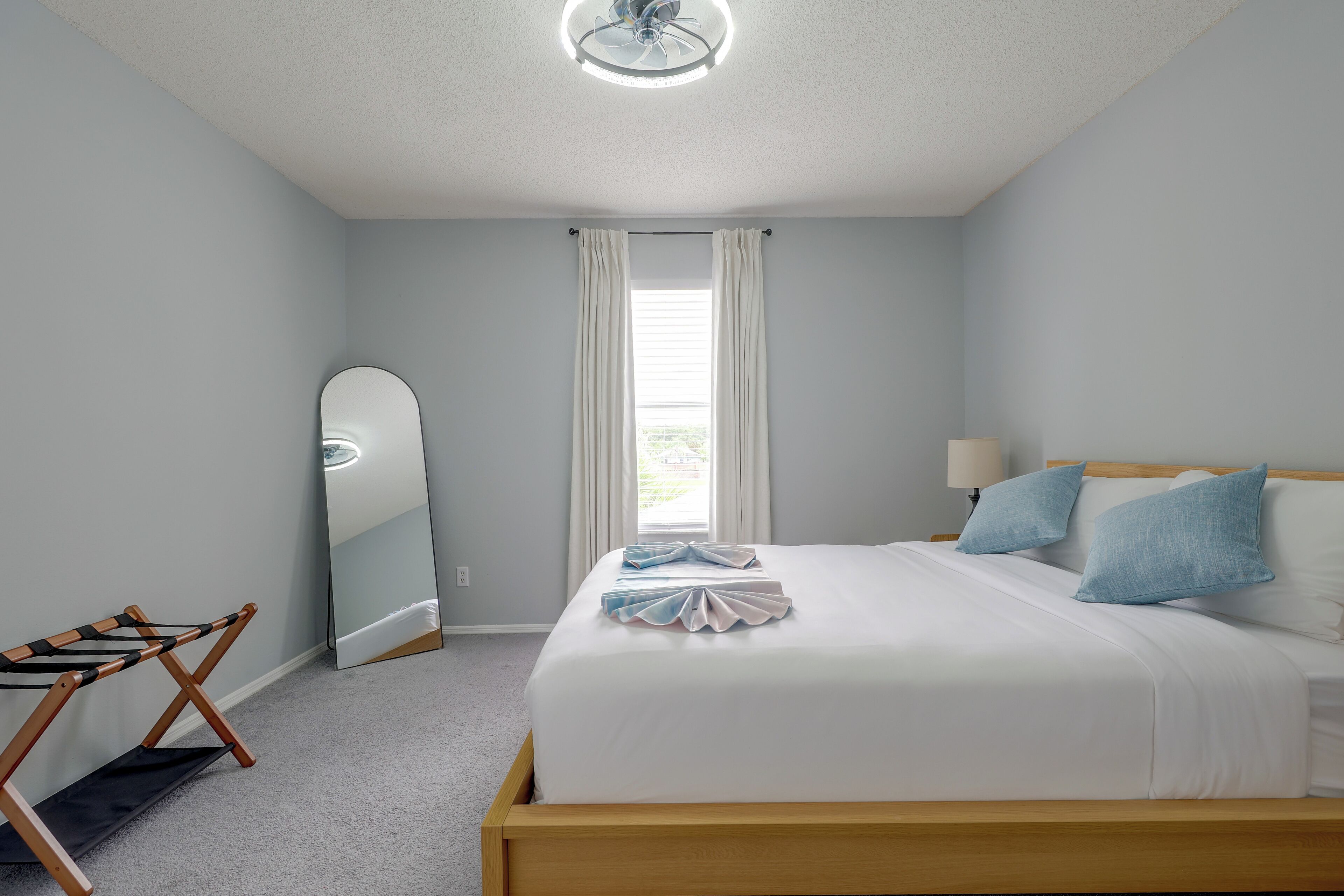 A bedroom with a bed, a mirror, a window with curtains, a lamp, and a wooden chair.