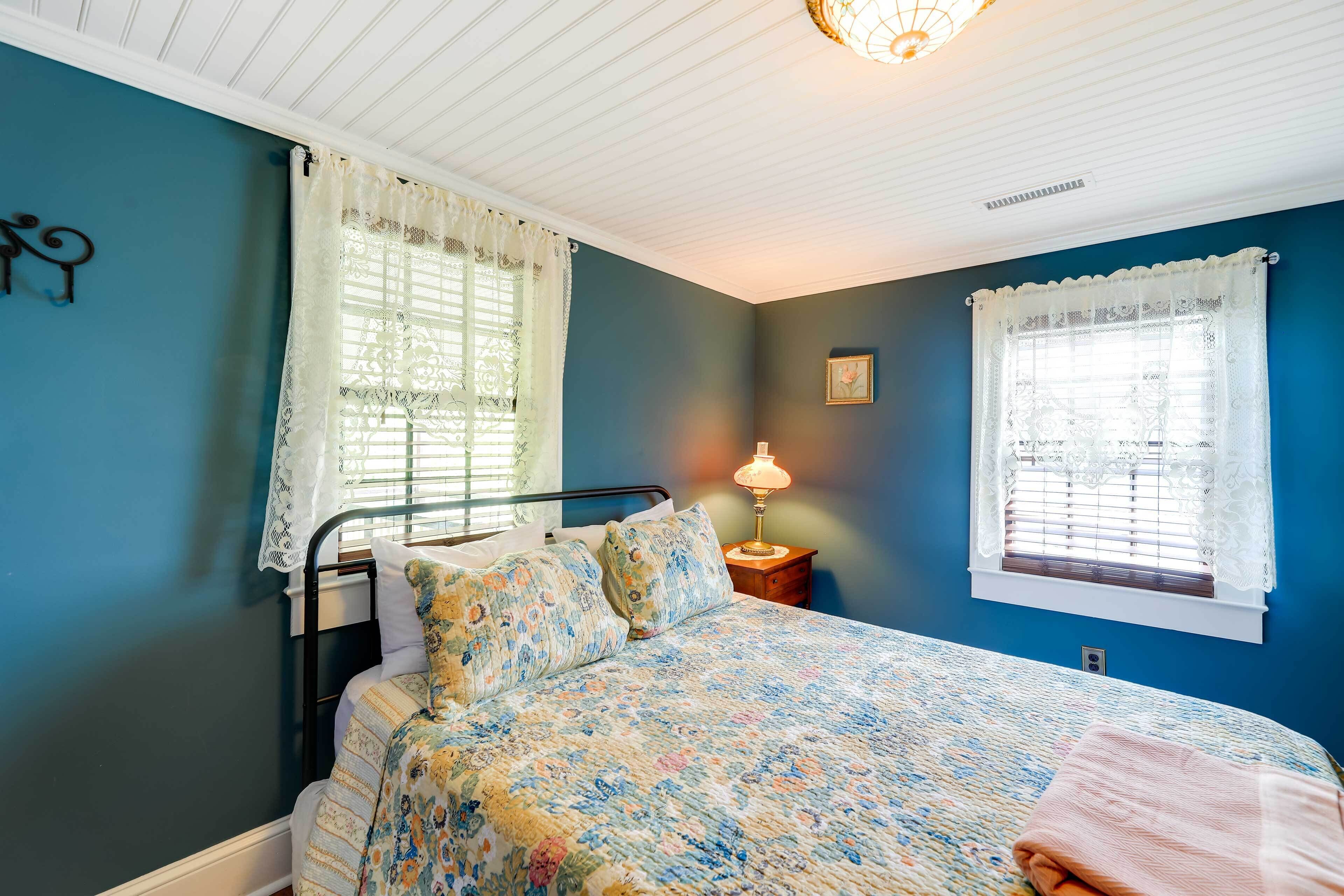 A bedroom with a bed, two windows with lace curtains, a bedside lamp, and a wall clock.