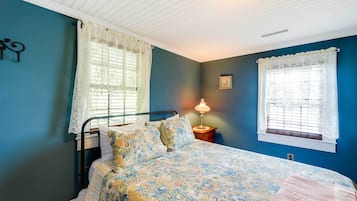A bedroom with a bed, two windows with lace curtains, a bedside lamp, and a wall clock.
