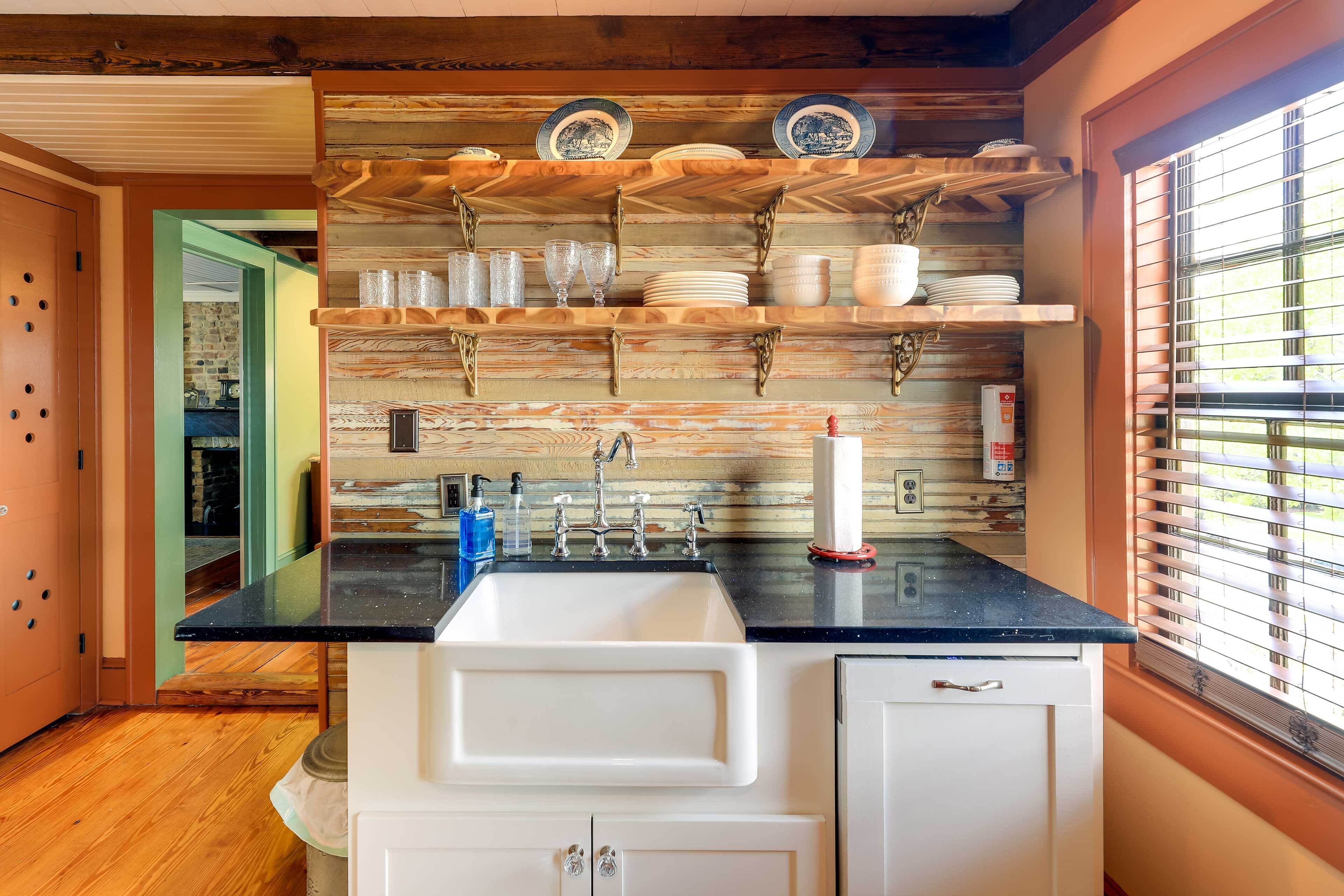 A kitchen with a wooden backsplash, a black countertop, and white cabinets. There are shelves above the sink holding plates and glasses.