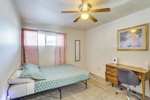 Un dormitorio con una cama individual, un escritorio con silla, un ventilador de techo, una ventana con cortinas y un cuadro en la pared.