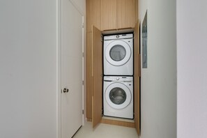 A compact laundry room with two stacked washing machines, a wooden cabinet, and a white door. - 4 Mi to Dtwn Mcallen, Sleek Couples Condo! (McAllen)