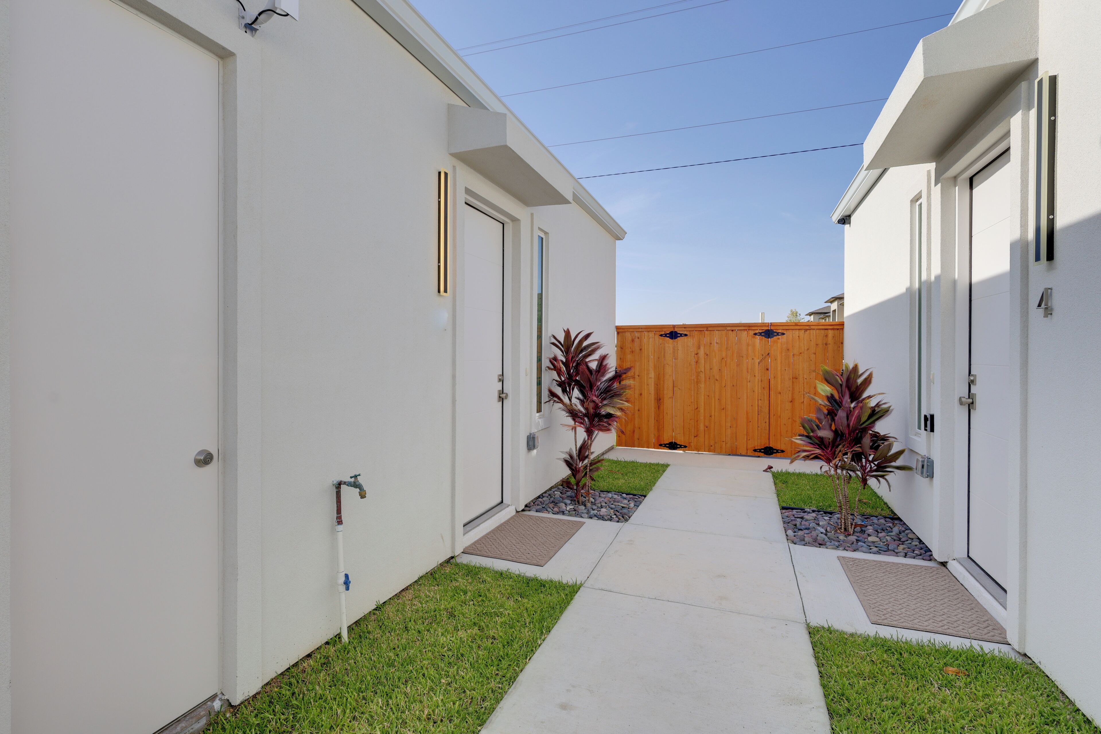 A narrow pathway leading to a wooden gate, flanked by white buildings with doors and windows, and small plants in the ground.