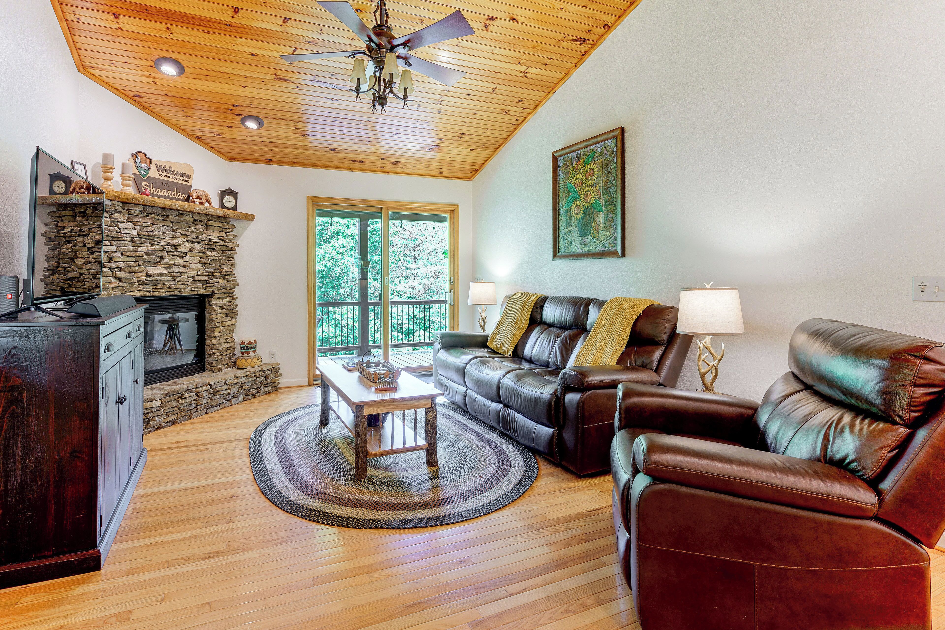 A living room with a stone fireplace, leather sofas, a wooden ceiling, and a large window.