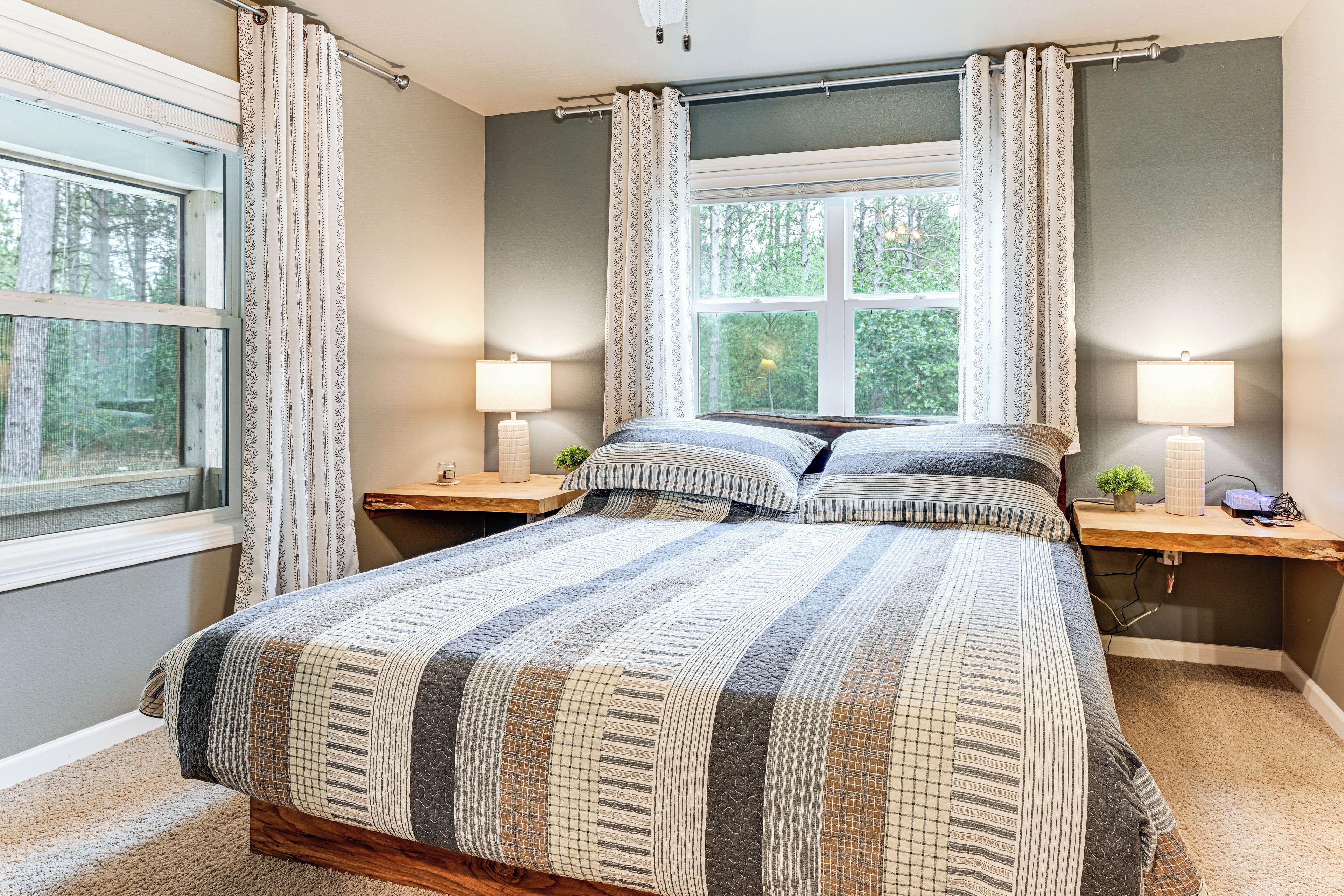 Un dormitorio con una cama grande, dos mesitas de noche, un ventanal con cortinas y vista a árboles.