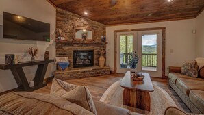 A cozy living room with a stone fireplace, a flat-screen TV, and a large window offering a view of the outdoors. - Blue Ridge Views, Hot Tub! Murphy Mtn Cabin (Murphy)