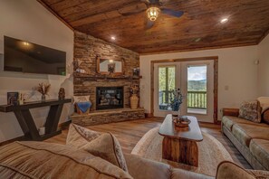 A cozy living room with a stone fireplace, a flat-screen TV, and a large window offering a view of the outdoors. - Blue Ridge Views, Hot Tub! Murphy Mtn Cabin (Murphy)