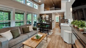 A spacious living area with a sofa, coffee table, and dining table. There is a kitchen area with a refrigerator and potted plants. - Tiny Home - Fire Pit, Grill, Deck & View! (Otto)