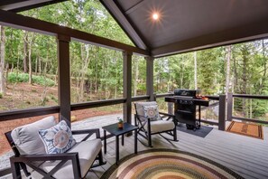A covered outdoor seating area with a grill, chairs, and a view of a forest. - Tiny Home - Fire Pit, Grill, Deck & View! (Franklin)