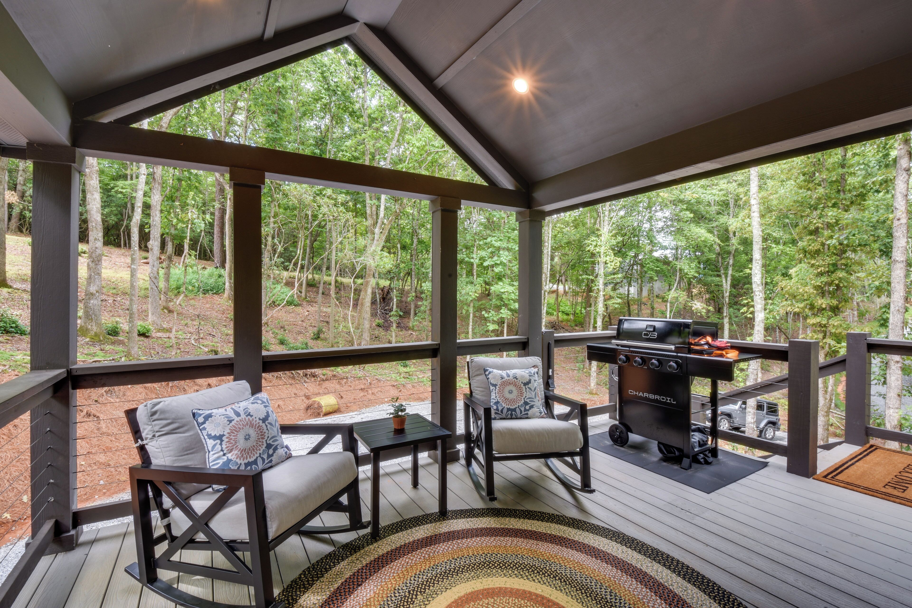 A covered outdoor seating area with a barbecue grill, cushioned chairs, and a view of a forest.