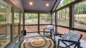 A covered porch with a rocking chair and a table, overlooking a forested area. - Tiny Home - Fire Pit, Grill, Deck & View! (Otto)