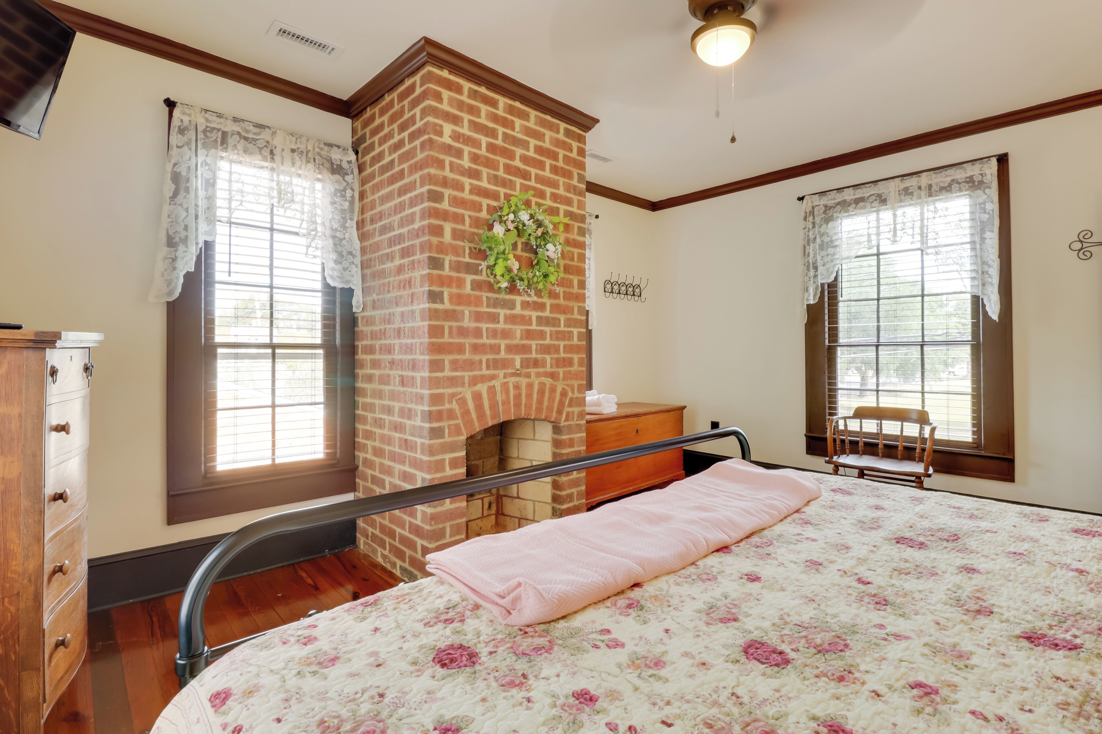 A bedroom with a brick fireplace, a bed with a floral bedspread, a wooden dresser, and two windows with lace curtains.