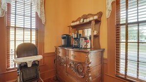 A corner room with a wooden dresser, a baby high chair, and a window with blinds.