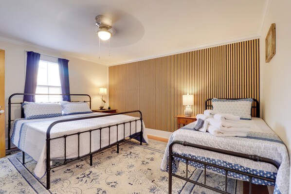 A bedroom with two beds, a window with curtains, a bedside table with a lamp, and a wall with vertical wooden slats.