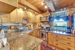 A kitchen with wooden cabinets, a granite countertop, a stove, a microwave, and hanging pots.