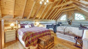 Une chambre à coucher rustique avec un plafond en bois, un lit recouvert d’une couette à carreaux, une table de chevet, une lampe, une commode en bois et une tête de cerf accrochée au mur.