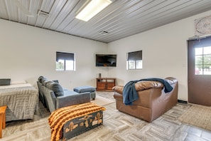 A spacious living room with a leather sofa, a couch, and a patterned ottoman. A flat-screen TV is mounted on the wall, and there are two windows providing natural light. - Montrose Studio Getaway w/ Covered Patio (Montrose)
