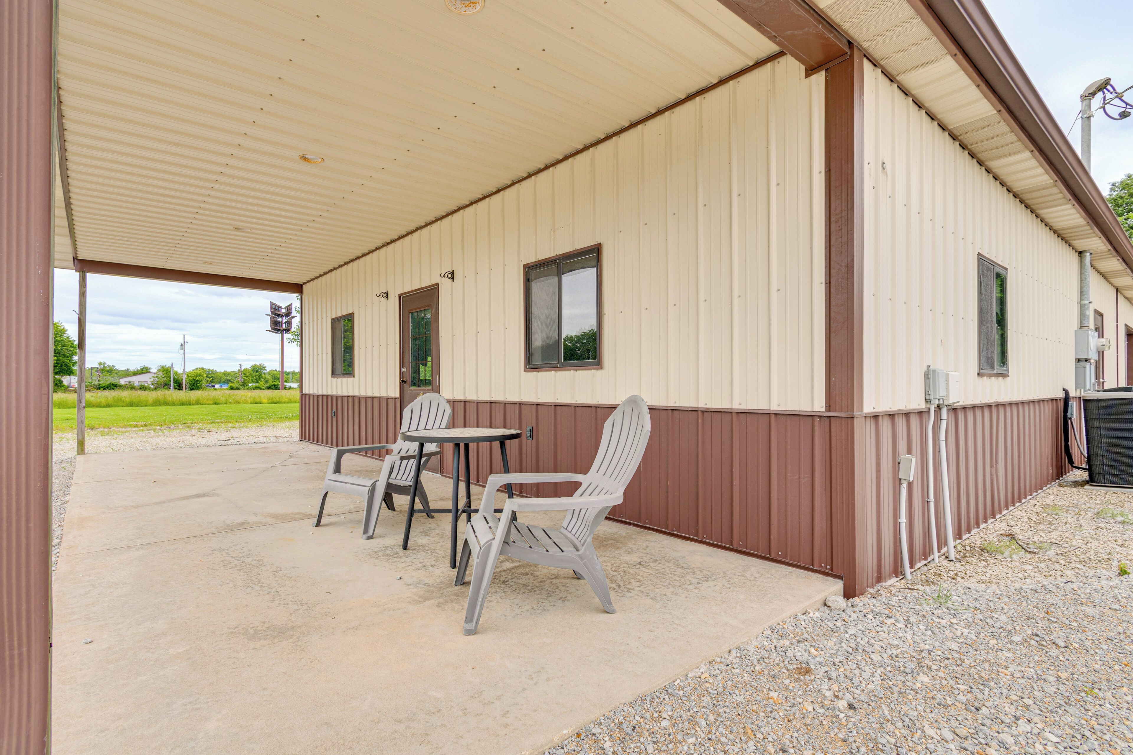 A covered outdoor area with a table and two chairs, a building with windows, and a gravel ground.