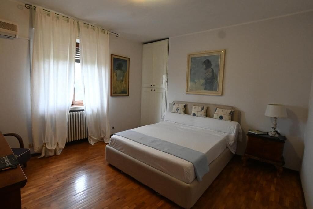 A bedroom with a bed, a radiator, a window with curtains, a nightstand, and a painting on the wall.