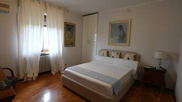 A bedroom with a bed, a radiator, a window with curtains, a nightstand, and a painting on the wall.