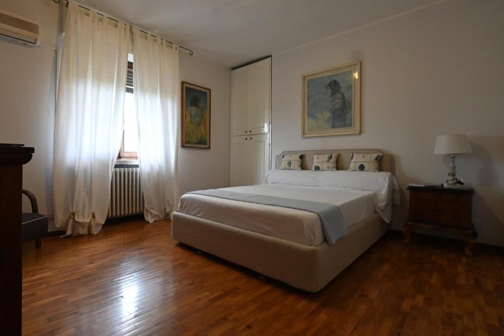 Quarto com cama, radiador, janela com cortinas, armário, pintura e mesa de cabeceira com abajur.