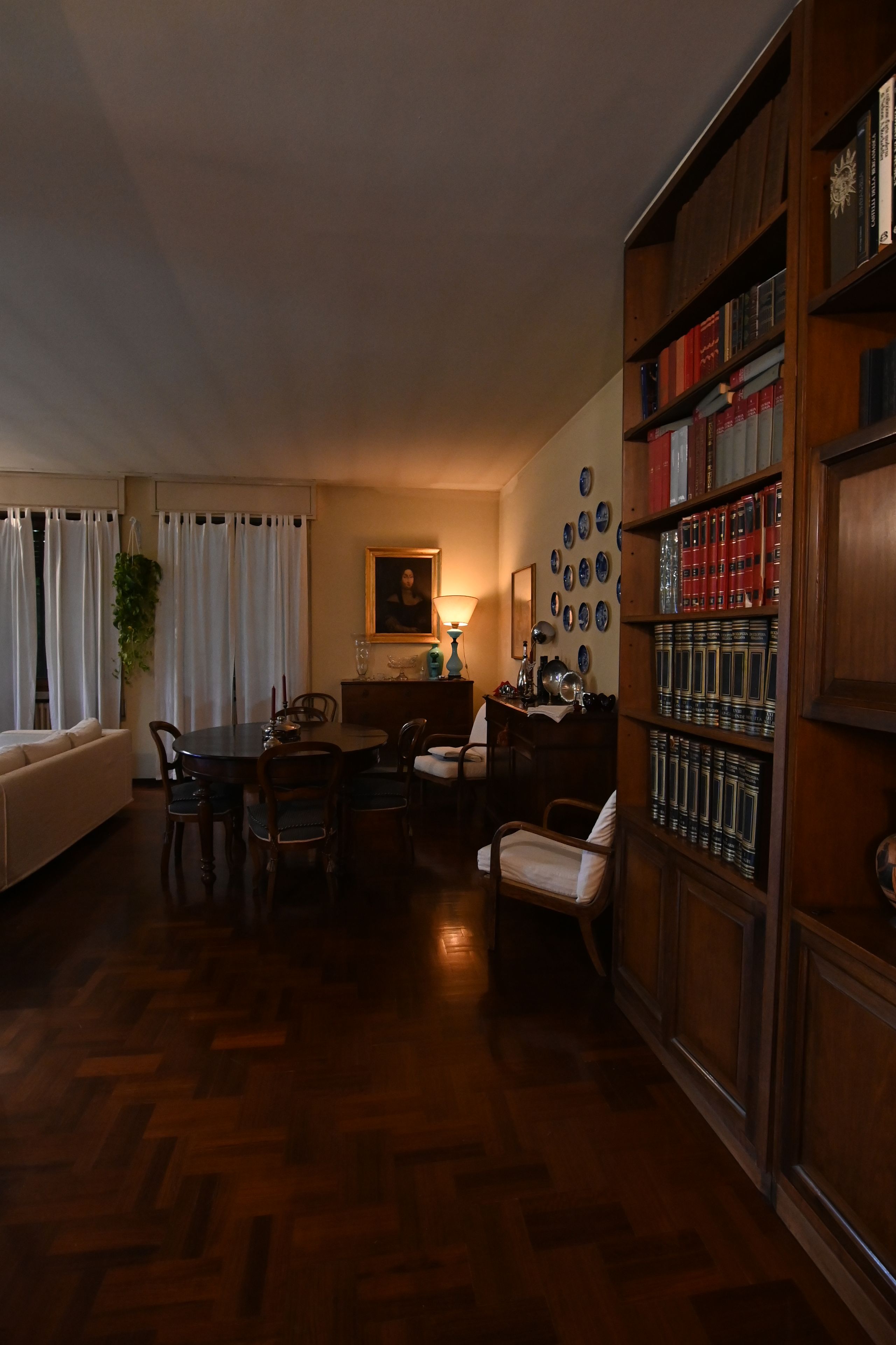 A room with a dining table, chairs, a bookshelf, a painting, and a window with curtains.