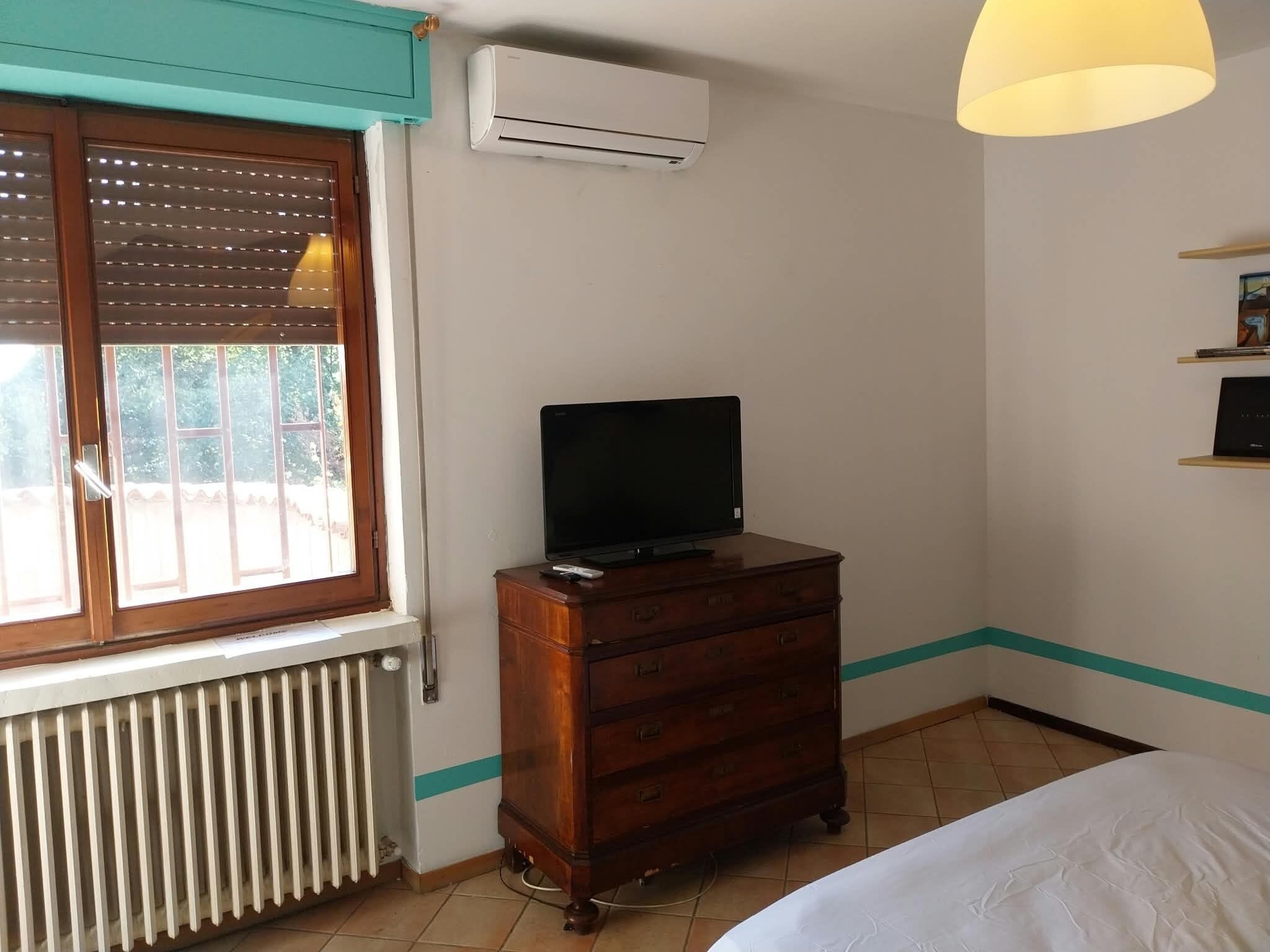 Ein Zimmer mit einem Bett, einem Holzschrank, einem Fernseher darauf, einer Klimaanlage, einem Fenster mit Jalousien und einem Heizkörper.