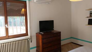 Ein Zimmer mit einem Bett, einem Holzschrank, einem Fernseher darauf, einer Klimaanlage, einem Fenster mit Jalousien und einem Heizkörper.