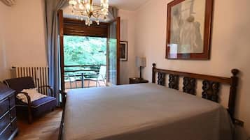 A bedroom with a large bed, a dresser, a chair, a table, and a view of greenery outside.