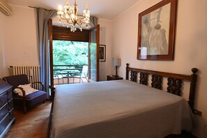 A bedroom with a large bed, a wooden headboard, a chandelier, a radiator, a chair, and a view of greenery outside.