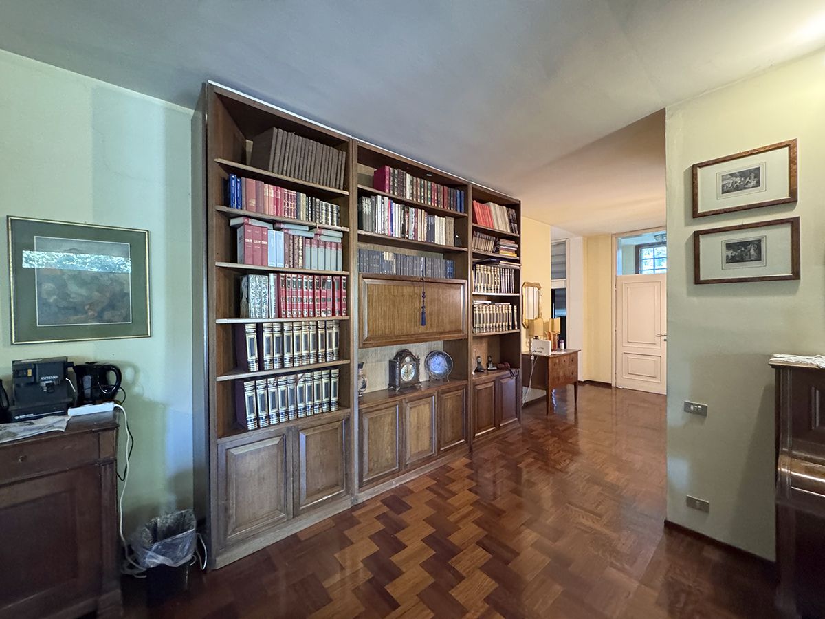 A spacious room with a large wooden bookshelf filled with books and decorative items, a wooden floor, and a door leading to another room.