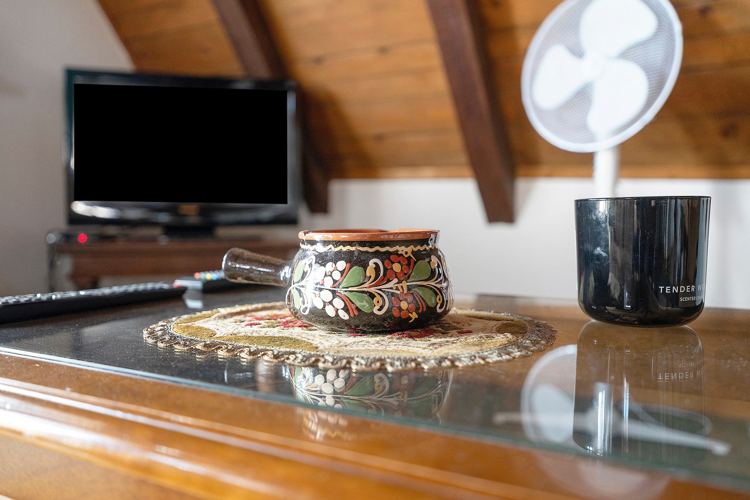 A glass table with a decorative mug and a black cup, a remote control, and a television in the background.