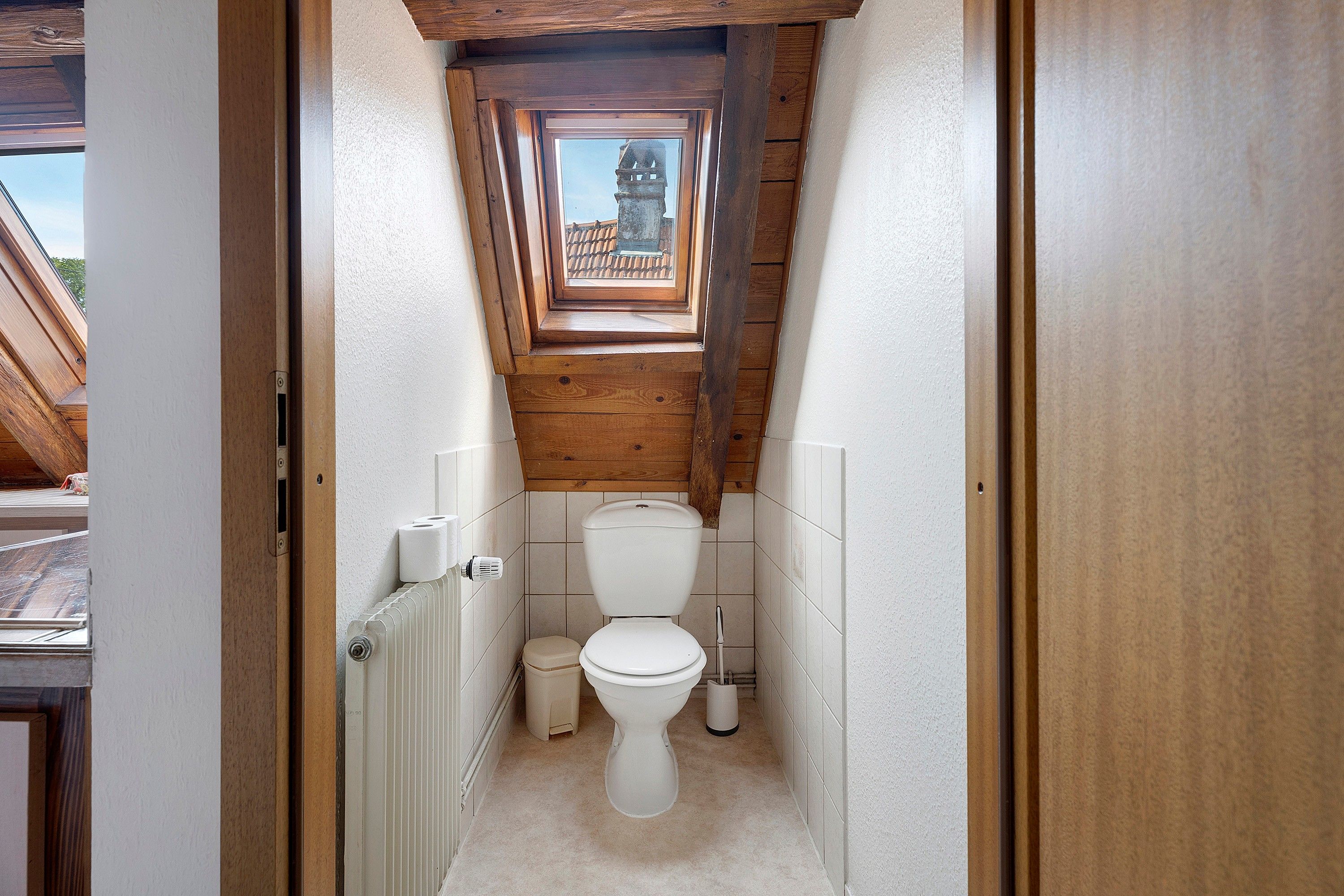A narrow, wooden-paneled bathroom with a skylight, white toilet, and a radiator.