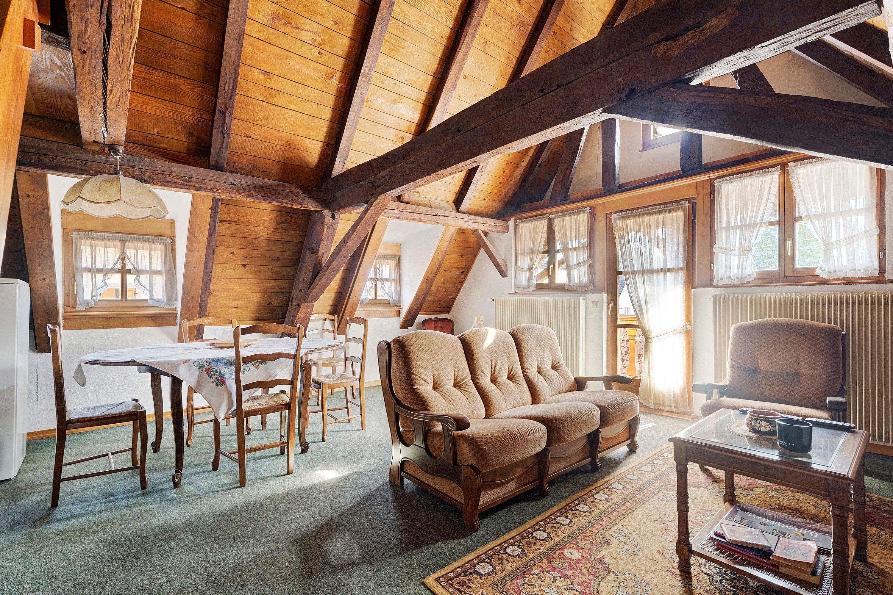 A cozy room with a sloped ceiling, wooden beams, a dining table with chairs, a sofa, and a coffee table with books and a mug.