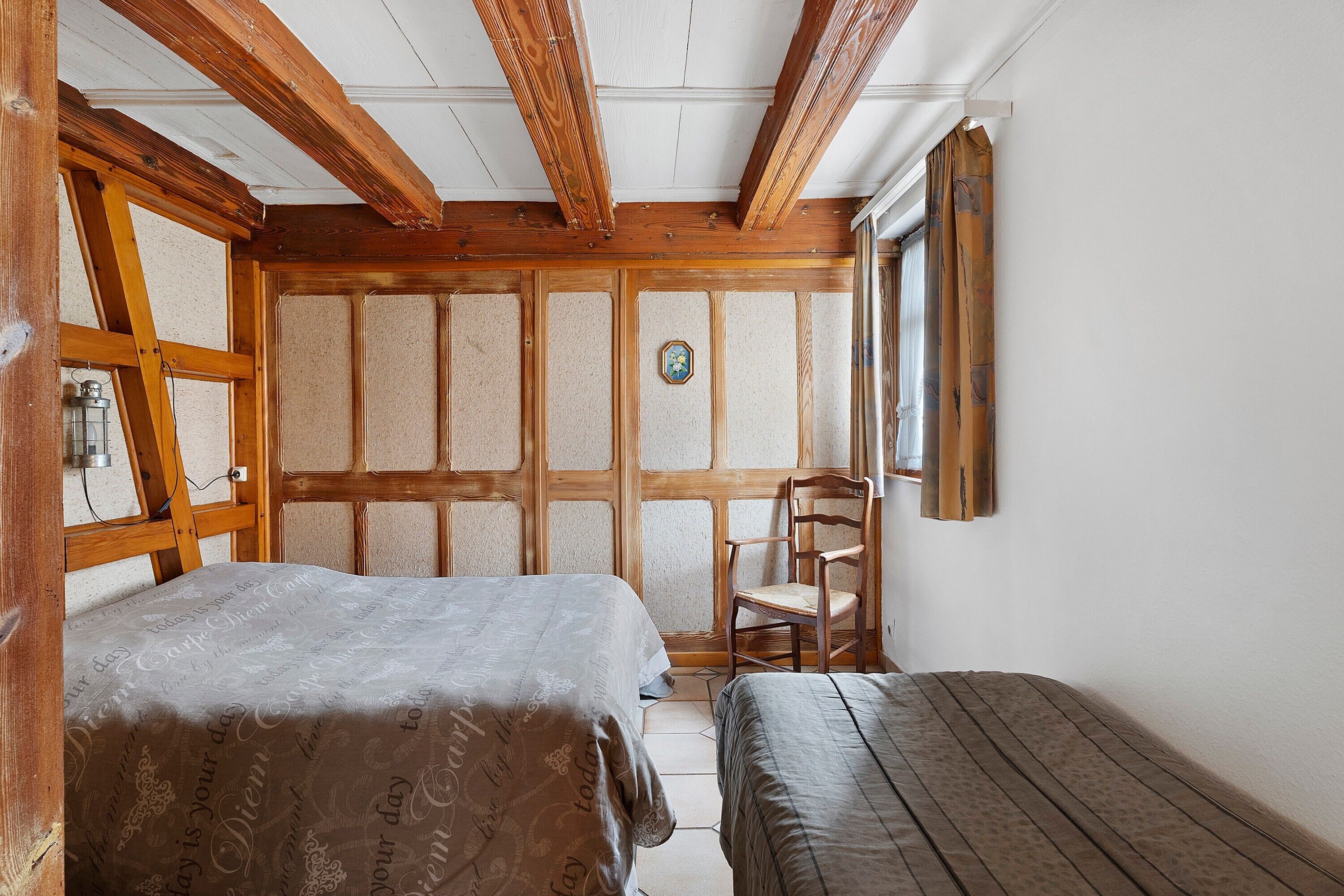 Ein Zimmer mit Holzbalkendecke, einem Bett, einem Sessel und einem Fenster mit Vorhängen.