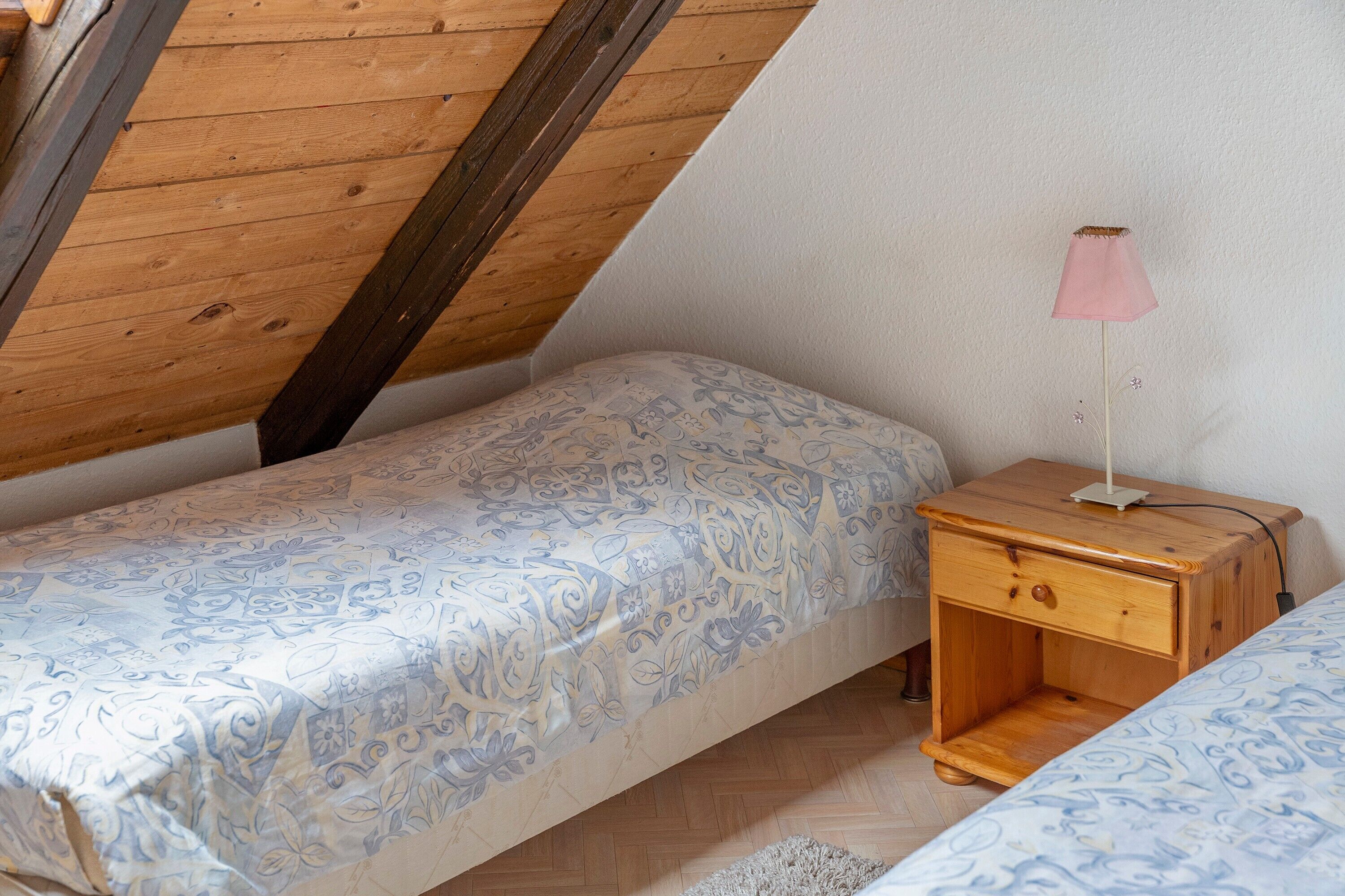 Een kleine, knusse kamer met een schuine kap, een enkel bed met beddengoed met een patroon, een nachtkastje met een lamp en een klein houten nachtkastje.