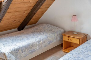 Habitació petita i acollidora amb sostre inclinat, un llit individual amb roba de llit estampada, una taula de nit amb una làmpada i una petita taula de nit de fusta.