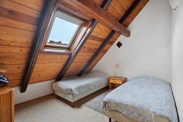 A small, sloped-roofed room with two beds, a wooden desk, and a window providing a view of the outside.