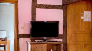 En tv-apparat står på ett träställ med ett skåp under, en trädörr finns till höger, och väggen är rosa med en träliknande struktur.