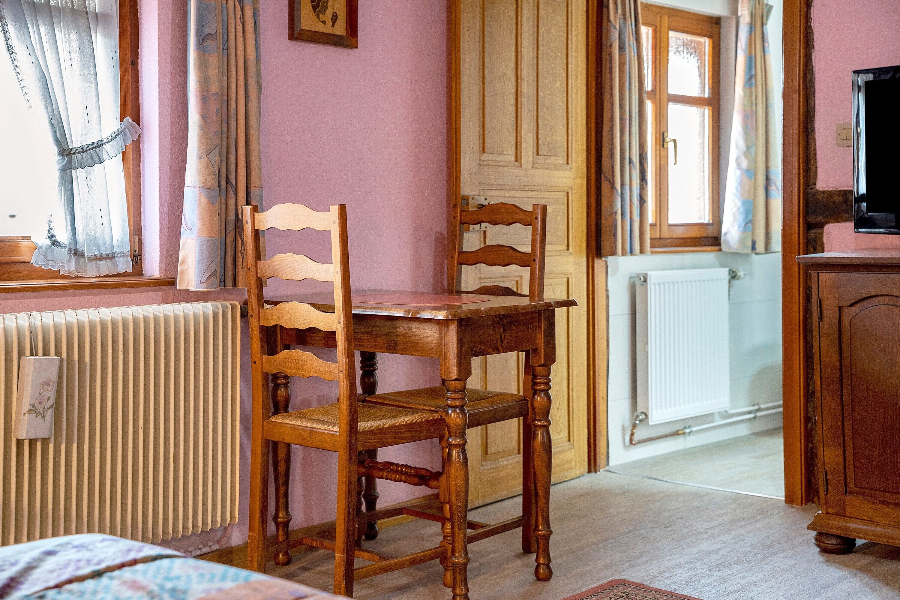 Um quarto com uma mesa e cadeiras de madeira, um radiador, uma janela com cortinas e uma porta.
