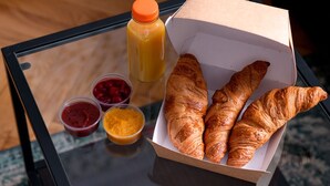 Una mesa de vidrio con una caja de cruasanes, un botellón de jugo de naranja y recipientes pequeños de mermelada y miel.