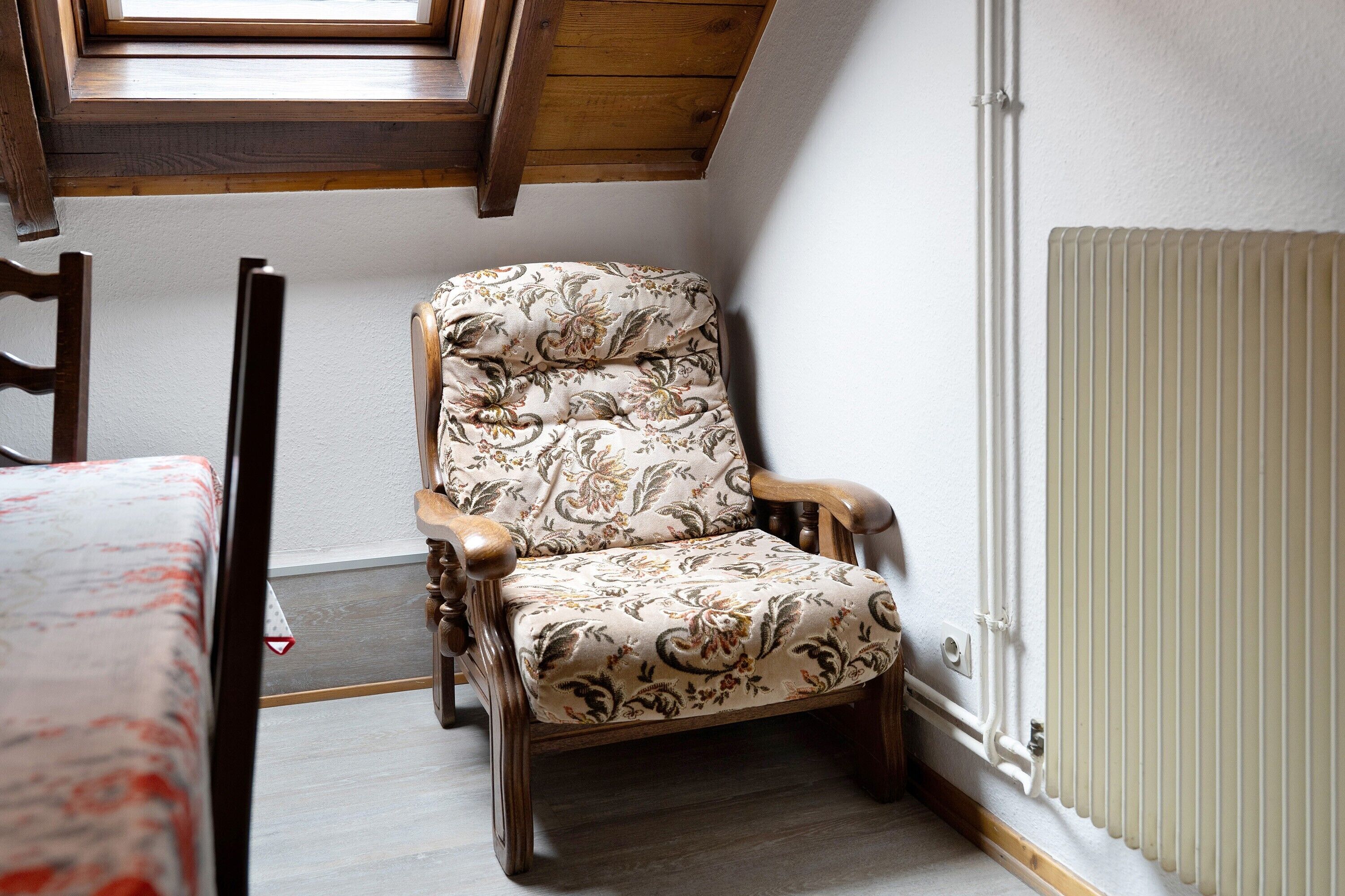 Une chaise en bois, recouverte d’un tissu à motifs floraux, se trouve dans une pièce dont le plafond est en pente et qui est équipée d’un radiateur.