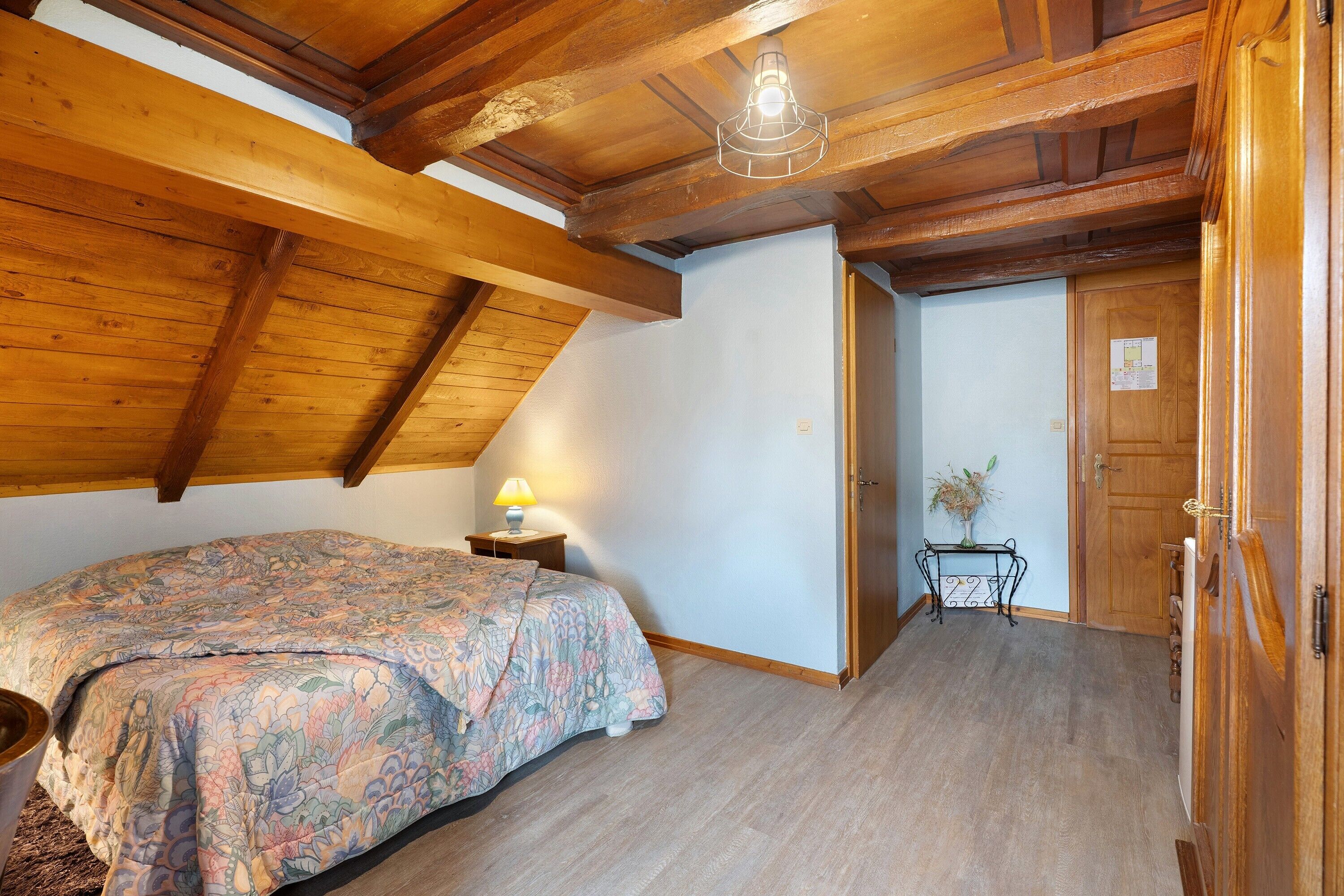 Ein Schlafzimmer mit schrägen Wänden, einem Bett mit Blumenmuster auf der Bettdecke, einem Nachttisch mit Lampe und einer Tür zu einem weiteren Raum.